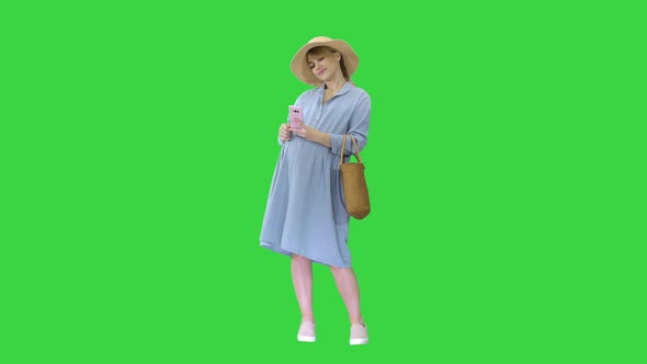 Happy Pregnant Woman in Blue Dress and Hat Using Her Smartphone on a Green Screen Chroma Key alt