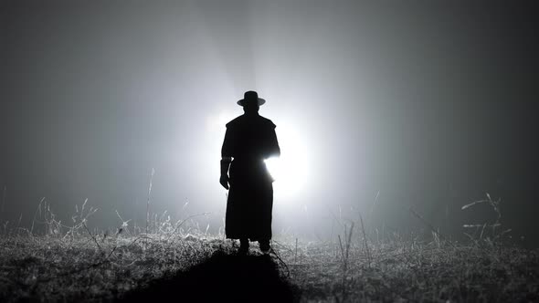 Silhouette of Man in Long Mantle Costume and Hat of Plague Doctor Is Walking Through Smoky Right alt