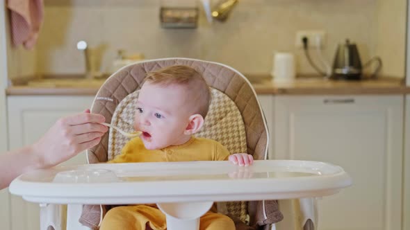 Mother feeding toddler baby with spoon on high chair for children, home kitchen background