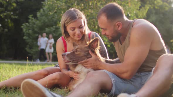 Young Couple Sitting in Park on Grass and Petting Their Dog on Sunny Summer Day