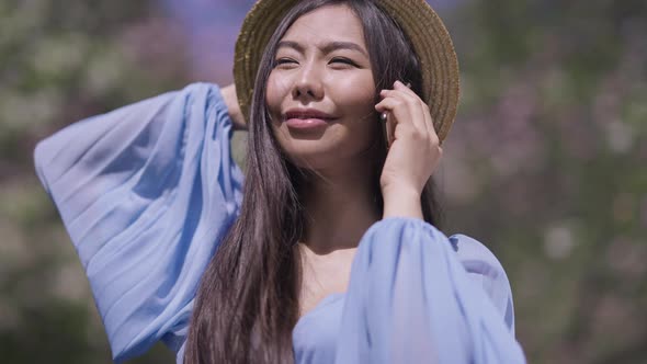 Positive Young Beautiful Asian Millennial Talking on Smartphone Smiling Standing in Sunny Sakura alt