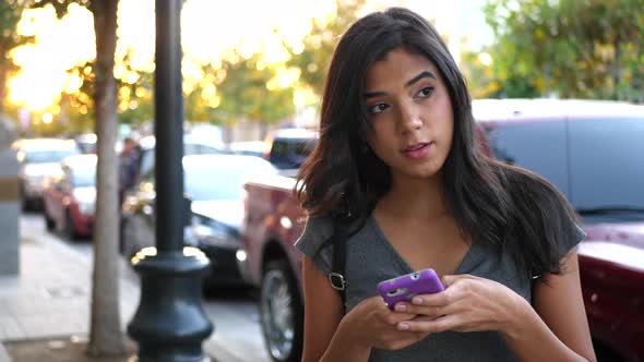 A young woman walking on an urban city street sidewalk texting or using social media on her smartpho alt