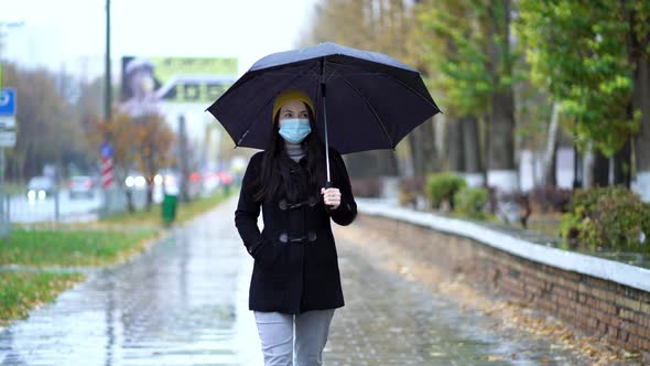 A Young Woman in a Protective Mask Walking in the Park Under Umbrella, Rainy Day, During Second Wave