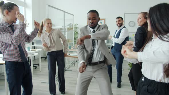 Joyful African American Man Dancing Among Multiethnic Group of Colleagues Having Fun in Workplace alt