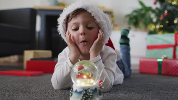 Surprised caucasian boy wearing santa hat lying on floor, watching snow globe alt