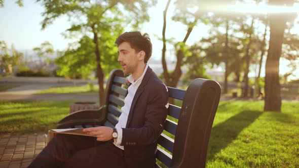 Slow Motion of Handsome Executive Business Man in Suit Sitting on Bench in Green Park Reading