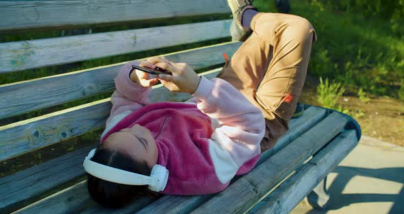 Ten Years Old Girl with Smartphone and in Headphones She Lying on the Bench and Chatting with alt