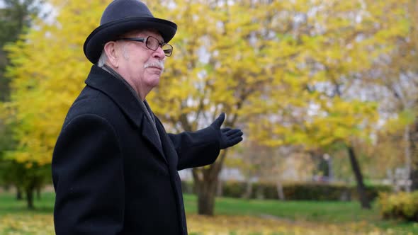 An Old Man Dressed in a Black Coat and Hat Tells the Story By Pointing His Hands to the Side alt
