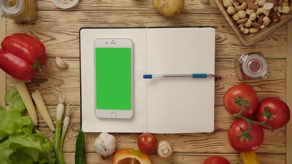Pen Rolling on Blank Notebook with Smartphone on Kitchen Table with Vegetables alt