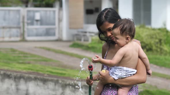 A Woman with a Child Not Far From the House Holds a Hose From Which a Stream of Water Pours and
