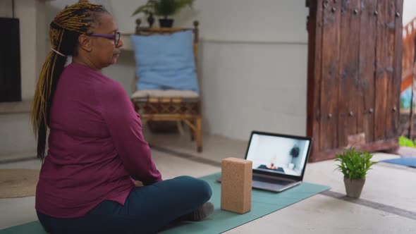 African senior woman doing yoga virtual fitness class with laptop at home alt
