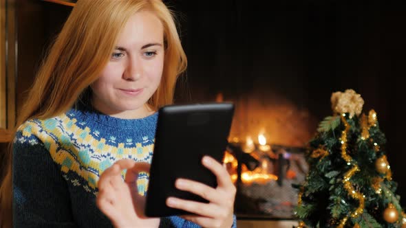A Young Woman Uses Tablet Near a Christmas Tree on a Background of Fire alt