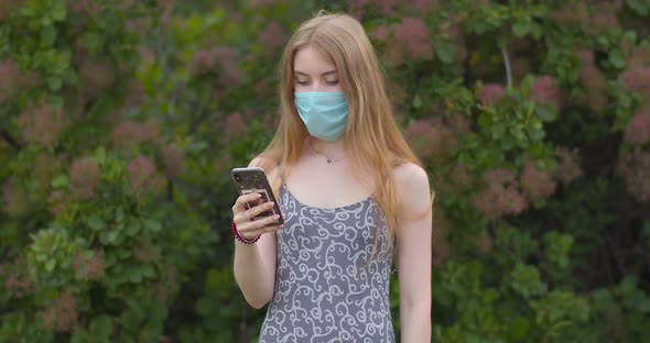 A young girl in a protective medical mask looks at the phone and sends messages