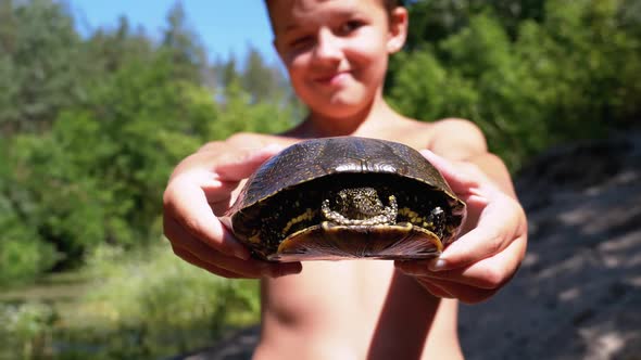 Boy Holds Turtle in Arms and Smiles on Background of River with Green Vegetation alt