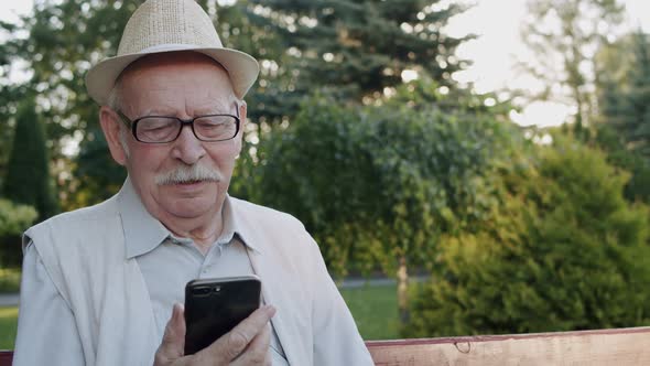 Senior Man in Hat and Suit Chatting on Phone's Web Camera in Park on Bench alt