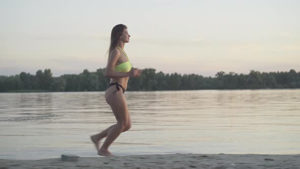 Side View of Slim Athletic Caucasian Woman Running Along Sandy Beach at Sunset. Wide Shot Portrait alt
