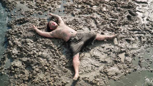 Funny Fat Man with Long Hair Lies and makes an Mud Angel. Overhead shot alt