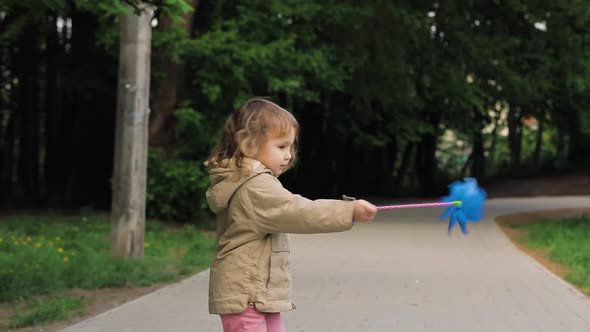 Cute Light Hair Little Girl with Windmill Toy Turns Around Herself. Girl Smiles and Plays, Outdoor