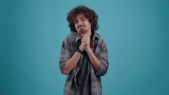 Young Hipster Asks for Help Praying with His Hands Together in the Studio on a Turquoise Background alt