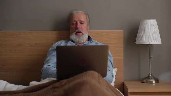 Front View of Grayhaired Senior Male with Beard Using Laptop Computer Lying on Comfortable Bed at alt