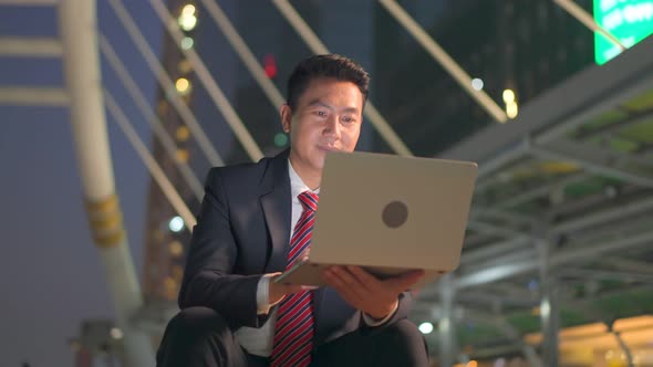 Asian businessman working on computer notebook sitting outdoor at night in city to complete job. alt