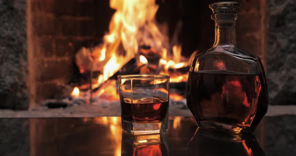 Bottle and Glass with Whiskey or Cognac on the Background of Fire in the Fireplace alt