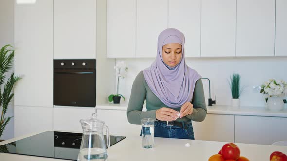 Progressive Arab Woman in a Hejab and Jeans Drinks Vitamins or Medicines in Her Kitchen alt