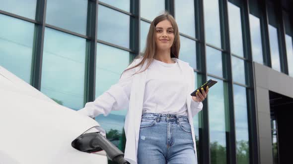 Young Woman is Standing Near the Electric Car and Looks at the Smart Phone alt