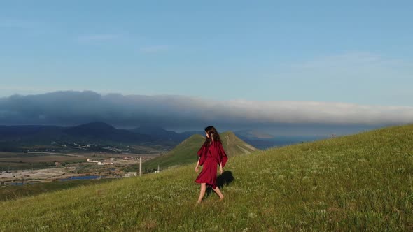 Side View of a Young Woman in a Red Dress That Flutters in the Wind Walking Along the Side of a Hill alt