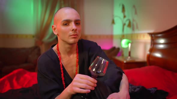 Depressed Male Queer Holding Wine Glass in Hand Thinking Sitting on Bed in Red Light alt