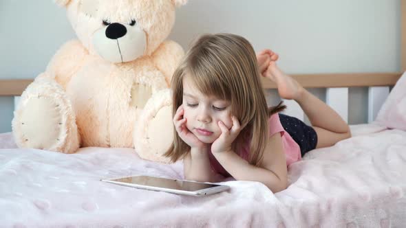 Little Child Girl Lies at Home in Bed and Watches Digital Tablet