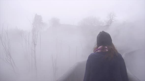 A young woman wearing a beanie hat, sweater and scarf hiking on a trail path in the fog. alt