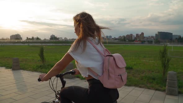 Beautiful young woman rides a bicycle in a city park during sunset alt
