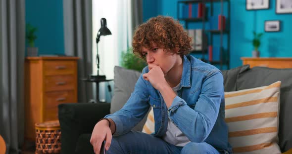 View of a Bored Teenager Sitting on a Sofa and Watching Television Switches Channels with alt