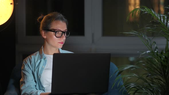 Woman with Glasses is Sitting in the Armchair and Working on a Laptop at Night alt