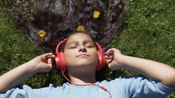 A Girl with Long Hair Lies on the Grass in the Park and Listens to Music in Headphones alt