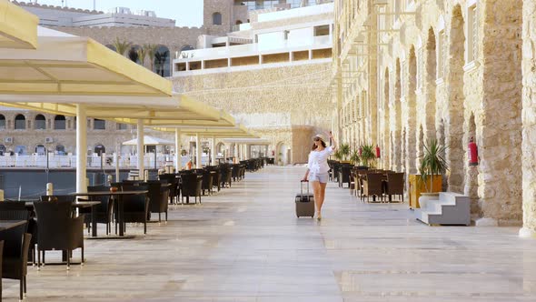 Young Woman in Summer Clothes, Sunglasses and Sun Hat, Walks with Travel Luggage Along a Deserted alt