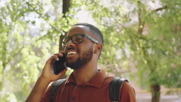 Cheerful Afro-American Man Walking in Park and Chatting on Phone alt