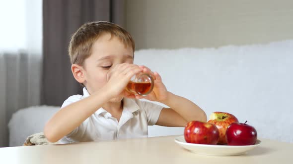 A Little Boy Is Sitting at a Table. A Glass of Fruit Juice and Apples Are on the Table. The Child alt