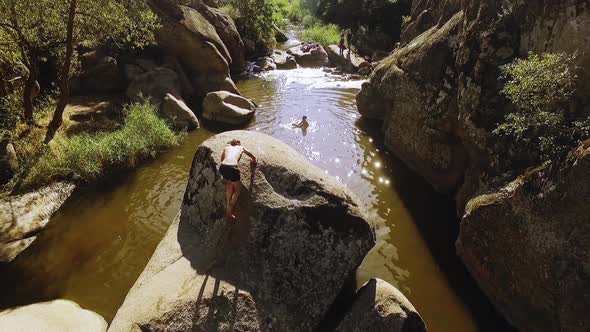 Male Climbing Big Stone in River Raising Tattooed Hands in Front of Friends alt