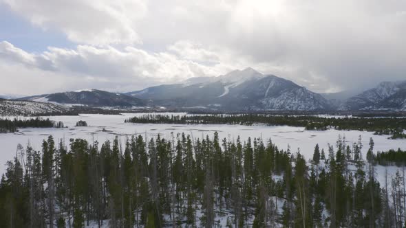 Aerial moving forward to reveal icy, frozen lake surrounded by snow and green pine trees with large alt
