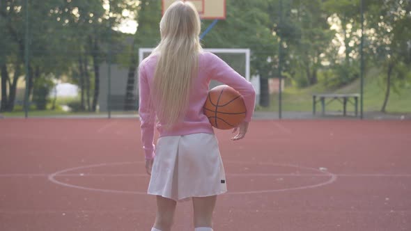 Back View Wide Shot of Sportive Blond Woman Standing with Basket Ball on Outdoor Court. Zoom in To alt