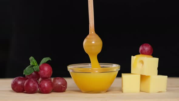 Dips wooden stick to glass bowl with liquid honey. Cheese, Honey and Grapes alt