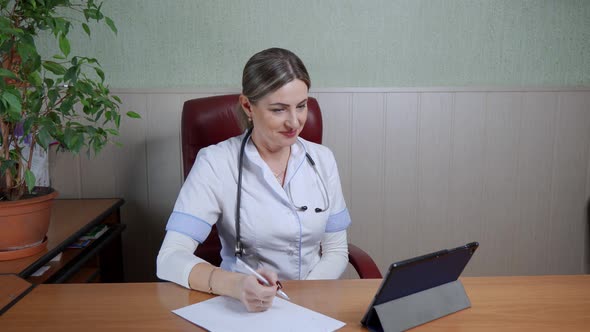 Smiling female doctor in a white coat gives a remote consultation to a patient using a tablet.