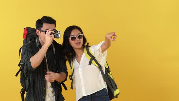 Smiling Asian backpacker couple taking photo follow woman pointing isolated on yellow background
