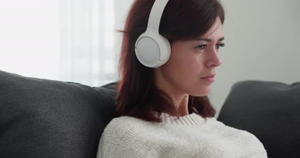 Portrait of Beautiful Woman Listening Music in Wireless Headphones While Relaxing on Grey Couch alt
