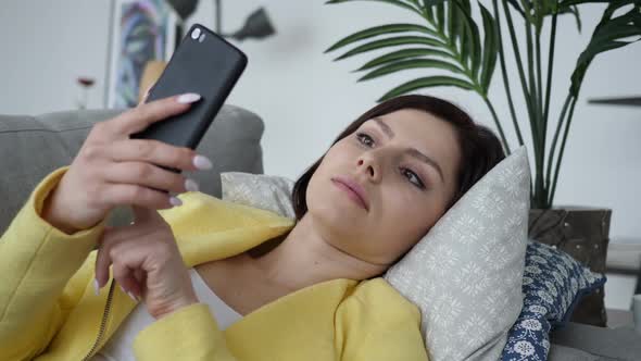 Woman Using Smartphone in Shock Wondering While Laying on Sofa alt