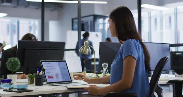 Caucasian businesswoman sitting at desk using laptop and writing notes in notebook in busy office alt