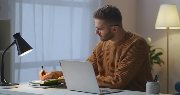 Adult Man Is Writing Notes in Notebook and Typing at Laptop Sitting Alone in Room Working Remotely alt