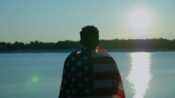 Afroamerican Man with American Flag on Shoulders Looks Into Distance at Sunrise alt